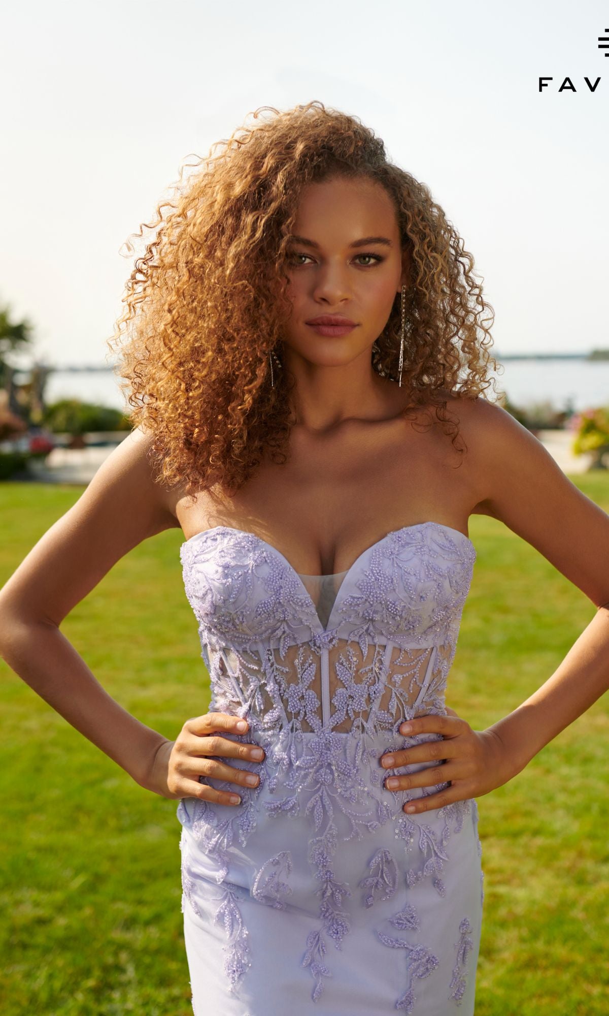 A woman with curly hair wears the Lilac Purple Strapless Faviana Long Prom Dress, outdoors on grass with water and trees in the background.